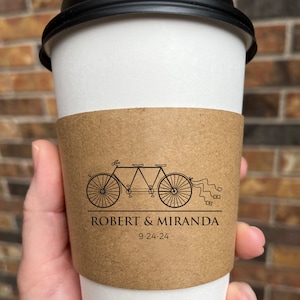 May include: A white paper coffee cup with a black lid and a brown sleeve. The sleeve features a tandem bicycle graphic and the names "ROBERT & MIRANDA" with the date "9-24-24".