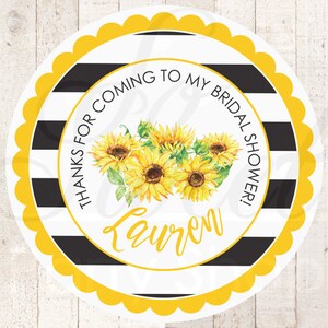 May include: Round, white favour tag with black and white stripes, a yellow scalloped border, and a sunflower design. The text reads "Thanks for coming to my bridal shower!" with the name "Lauren" below the sunflowers.