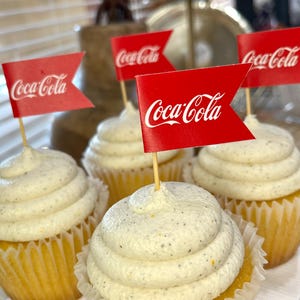 May include: Several cupcakes with white frosting and red Coca-Cola flags. The cupcakes are in yellow paper liners. The frosting has dark specks. The Coca-Cola flags are red with white text.
