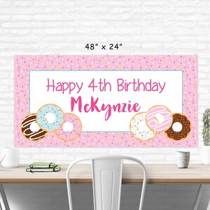 Op de afbeelding: Roze en witte verjaardagsbord met de tekst "Happy 4th Birthday Mckynzie" en donut-illustraties. De banner is 122 cm breed en 61 cm hoog.