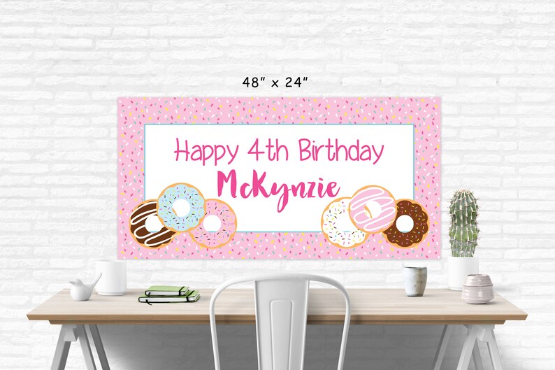 Op de afbeelding: Roze en witte verjaardagsbord met de tekst "Happy 4th Birthday Mckynzie" en donut-illustraties. De banner is 122 cm breed en 61 cm hoog.