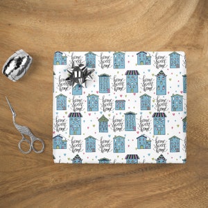 May include: White wrapping paper with a repeating pattern of blue houses with pink and green accents. The houses have windows and the text "home sweet home" is written on each house. There are also small pink hearts scattered throughout the pattern.