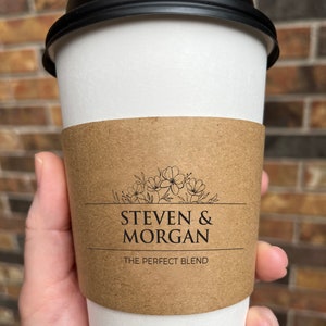 May include: A white paper coffee cup with a black lid and a brown sleeve. The sleeve features the text "STEVEN & MORGAN" above "THE PERFECT BLEND" with a floral design. The cup is held by a hand.