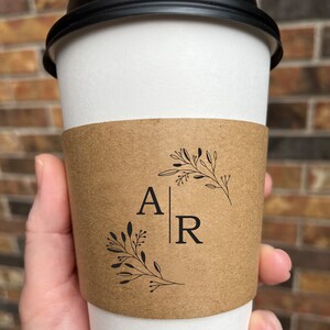 May include: A white paper coffee cup with a black lid. The cup is held by a hand and has a brown sleeve with the letters "A" and "R" separated by a vertical line, and floral designs.