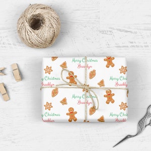 Op de afbeelding: Wit inpakpapier met een herhalend patroon van gingerbreadmen, bellen en sterren. Het papier heeft de tekst "Merry Christmas Brooklyn" die zich over de hele oppervlakte herhaalt.