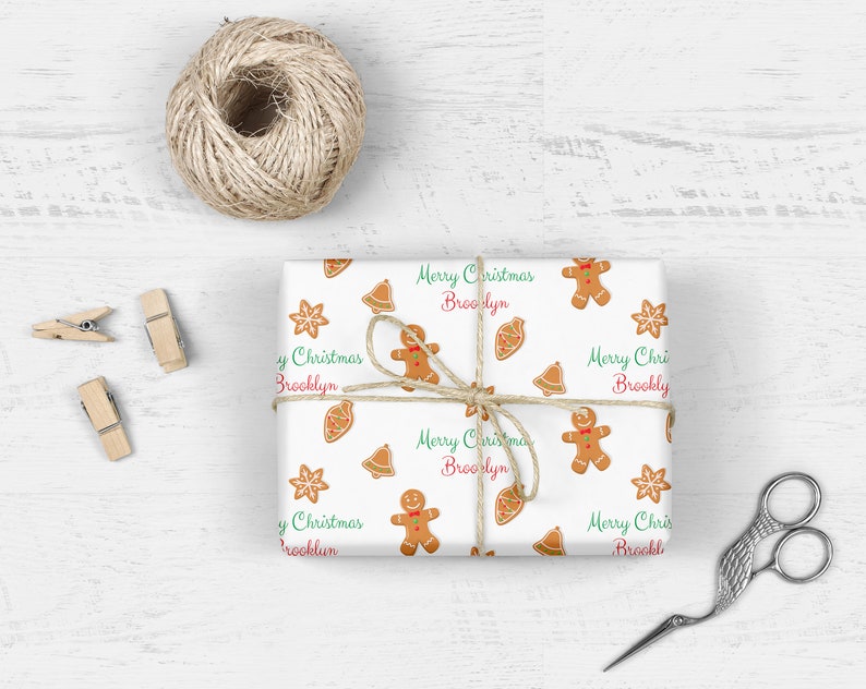 Op de afbeelding: Wit inpakpapier met een herhalend patroon van gingerbreadmen, bellen en sterren. Het papier heeft de tekst "Merry Christmas Brooklyn" die zich over de hele oppervlakte herhaalt.