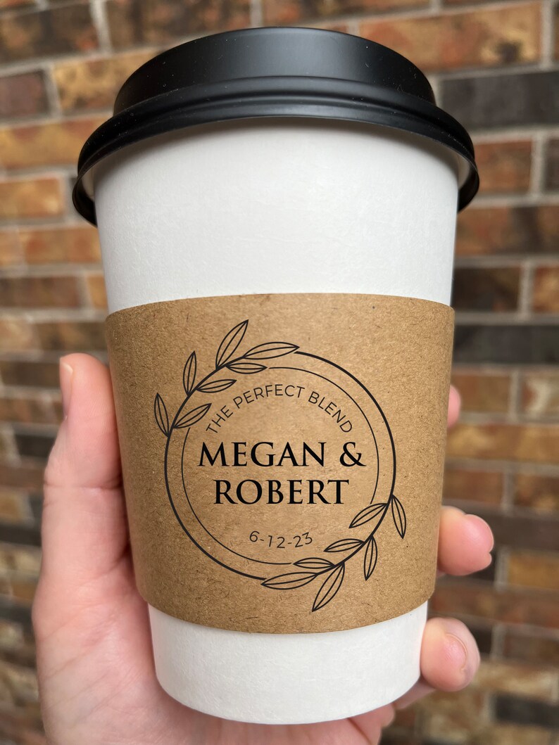 May include: A white paper coffee cup with a black lid. The cup has a brown sleeve with the text "THE PERFECT BLEND" and the names "MEGAN & ROBERT" with the date "6-12-23" inside a circular design with leaves.