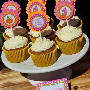 May include: A white cake with cream cheese frosting and a pumpkin-themed topper that says "Happy 1st Birthday Claudia". The cupcakes are on a white cake stand and there is a sign that says "Cream Cheese Cupcakes".