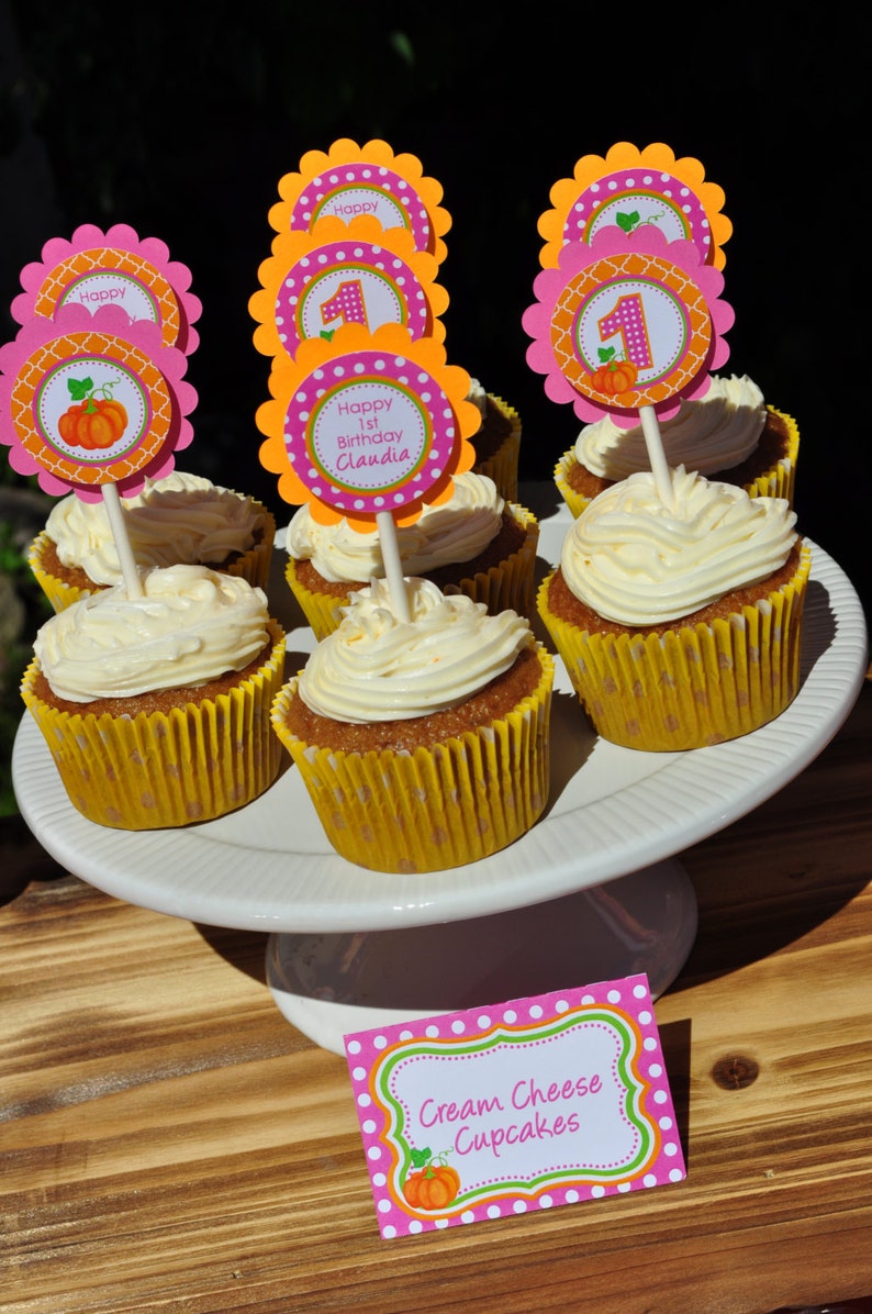 May include: A white cake with cream cheese frosting and a pumpkin-themed topper that says "Happy 1st Birthday Claudia". The cupcakes are on a white cake stand and there is a sign that says "Cream Cheese Cupcakes".