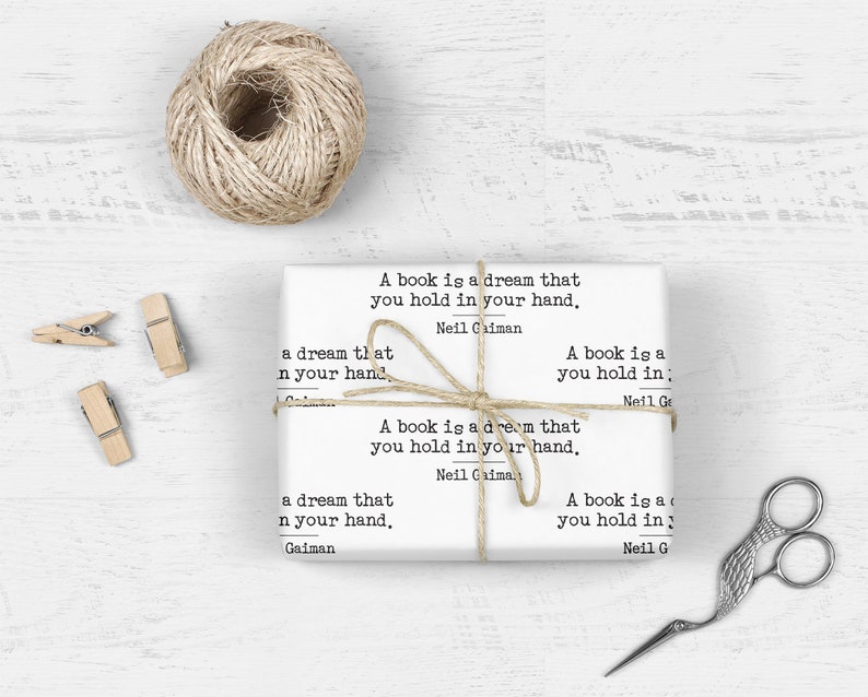 May include: White wrapping paper with the quote "A book is a dream that you hold in your hand" by Neil Gaiman. The paper is tied with twine and includes a ball of twine, wooden clothespins, and decorative scissors.