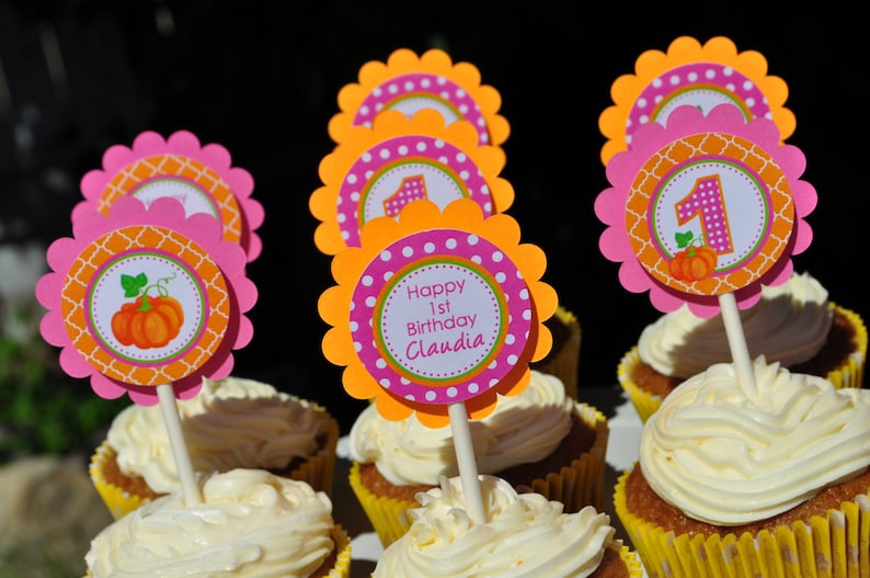 May include: Pink and orange cupcake toppers with a pumpkin design and the text "Happy 1st Birthday Claudia".