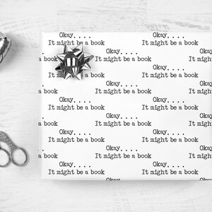 May include: White wrapping paper with the repeated text "Okay... It might be a book" printed on it. A silver bow is on the paper.