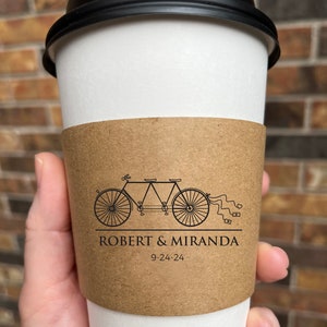 May include: A white paper coffee cup with a black lid, featuring a brown sleeve. The sleeve displays a tandem bicycle graphic with the names "ROBERT & MIRANDA" and the date "9-24-24".