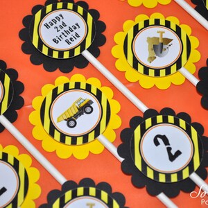 May include: Construction themed cupcake toppers with black and yellow striped circles. The toppers feature a dump truck, a shovel, a construction cone, and the number 2. The text "Happy 2nd Birthday Reid" is on one of the toppers.