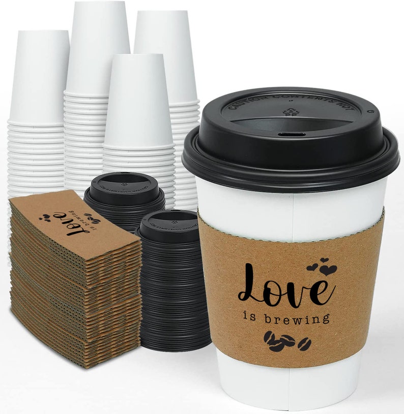 May include: A selection of white paper coffee cups, black lids, and cardboard sleeves. The sleeve on the front cup says "Love is brewing" with coffee bean and heart graphics. Stacks of cups and sleeves are visible.