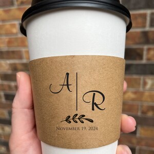 May include: A white paper coffee cup with a black lid, held by a hand. The cup has a brown sleeve with the initials "A" and "R" separated by a vertical line, with a leaf design and the date "November 19, 2024".