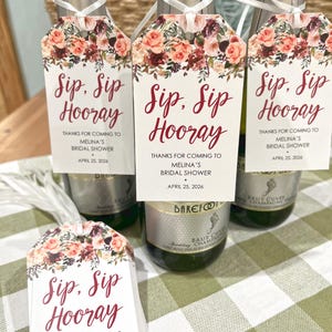 以下が含まれることがあります： ブルゴーニュ色の筆記体で「Sip, Sip, Hooray」と書かれたタグが付いたシャンパンボトル。花柄の装飾が施されています。タグには「Melina's Bridal Showerへお越しいただきありがとうございます。2026年4月25日」というテキストも含まれています。