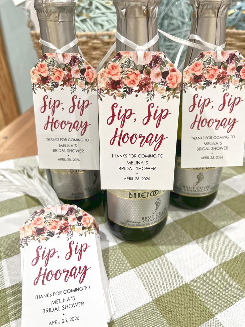 以下が含まれることがあります： ブルゴーニュ色の筆記体で「Sip, Sip, Hooray」と書かれたタグが付いたシャンパンボトル。花柄の装飾が施されています。タグには「Melina's Bridal Showerへお越しいただきありがとうございます。2026年4月25日」というテキストも含まれています。