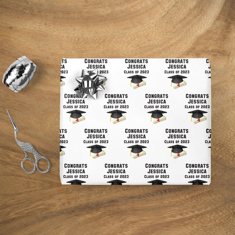 May include: White graduation-themed wrapping paper with a repeating pattern of graduation caps, diplomas, and the text "CONGRATS JESSICA CLASS OF 2023." A silver bow, scissors, and a roll of silver ribbon are on a wooden surface.