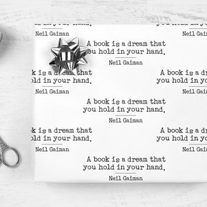 May include: White wrapping paper printed with the quote "A book is a dream that you hold in your hand" by Neil Gaiman. A silver bow and decorative scissors are next to the wrapped gift.