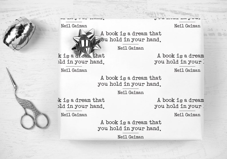 May include: White wrapping paper printed with the quote "A book is a dream that you hold in your hand" by Neil Gaiman. A silver bow and decorative scissors are next to the wrapped gift.