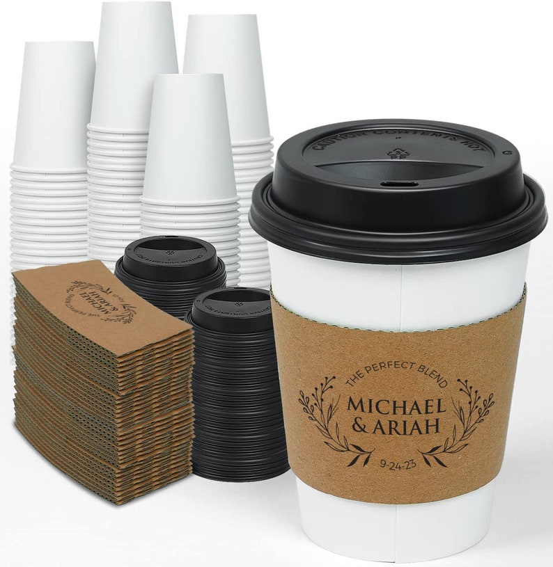 May include: White paper coffee cups with black lids, and brown cardboard sleeves. The sleeves are printed with the text "The Perfect Blend" and the names "Michael & Ariah" with a date. Stacks of cups and sleeves are visible.