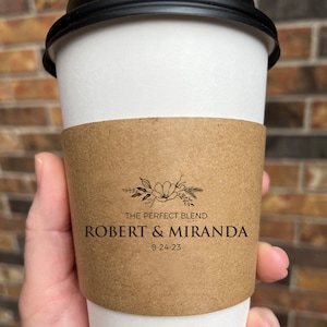 May include: A takeaway coffee cup with a black lid and a brown sleeve. The sleeve features the text "THE PERFECT BLEND", the names "ROBERT & MIRANDA", and the date "9-24-23". The cup is white.