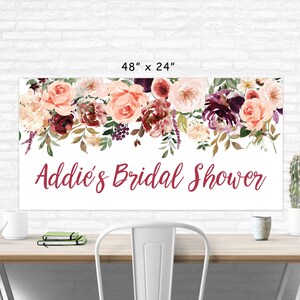 Op de afbeelding: Een witte bruidsdouchebanner met een bloemenkrans van roze, bordeauxrode en witte bloemen. De banner heeft de tekst "Addie's Bridal Shower" in bordeauxrode letters.
