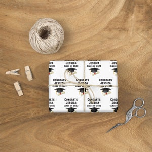 May include: White wrapping paper with a repeating pattern of graduation caps, diplomas, and the text "Congrats Jessica Class of 2023." The paper is tied with twine and rests on a wooden surface, with scissors and clothespins nearby.