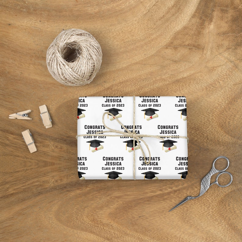 May include: White wrapping paper with a repeating pattern of graduation caps, diplomas, and the text "Congrats Jessica Class of 2023." The paper is tied with twine and rests on a wooden surface, with scissors and clothespins nearby.