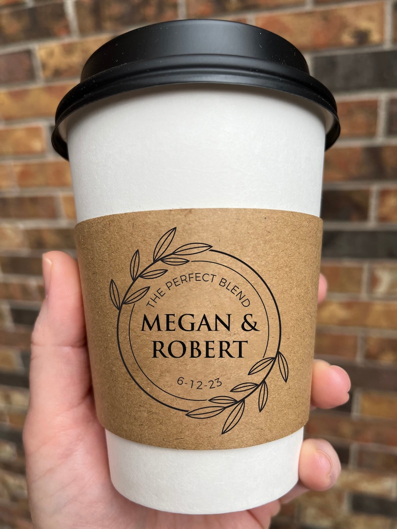 May include: A white paper coffee cup with a black lid. The cup is held by a person. A brown paper sleeve with the text "THE PERFECT BLEND" and the names "MEGAN & ROBERT" with the date "6-12-23" is wrapped around the cup. The background is a brick wall.