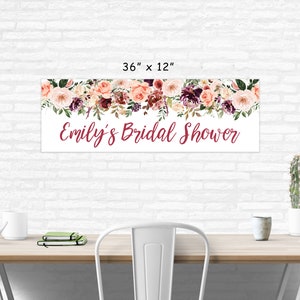 Op de afbeelding: Een witte banner met een bloemenkrans van roze, bordeauxrode en groene bloemen. De banner heeft de tekst "Emily's Bridal Shower" in bordeauxrode schrift. De banner is 91,44 cm breed en 30,48 cm hoog.