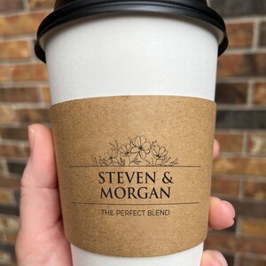 May include: A white paper coffee cup with a black lid and a brown sleeve. The sleeve has the text "STEVEN & MORGAN" above "THE PERFECT BLEND" with a floral design. The cup is held by a hand.