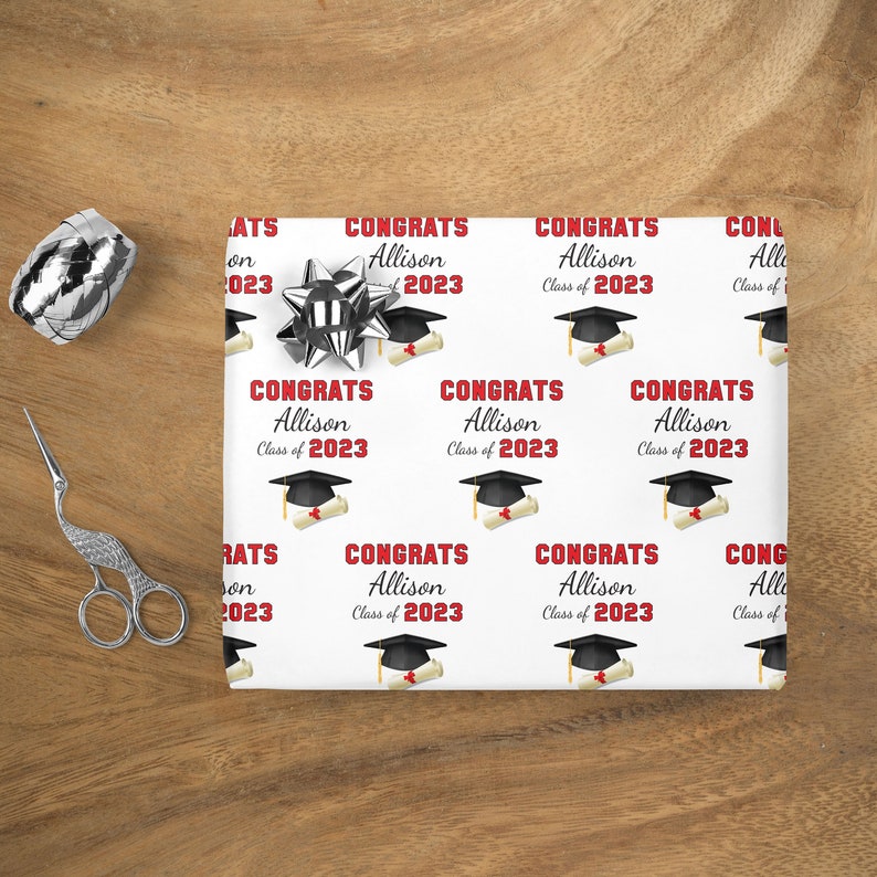 Op de afbeelding: Wit inpakpapier met een herhalend patroon van afstudeerhoeden, diploma's en de tekst "Congrats Allison Class of 2023" in rood.