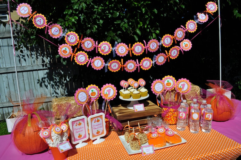 May include: A pink and orange pumpkin-themed birthday party table setting with a banner that reads "Happy Birthday Claudia". The table is decorated with cupcakes, candy, and a pumpkin centerpiece.