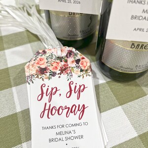 以下が含まれることがあります： 赤い筆記体で「Sip, Sip Hooray」と書かれた白いタグ。タグには「Melina's Bridal Showerへお越しいただきありがとうございます」と「2026年4月25日」と書かれています。背景にはシャンパンボトルがあります。