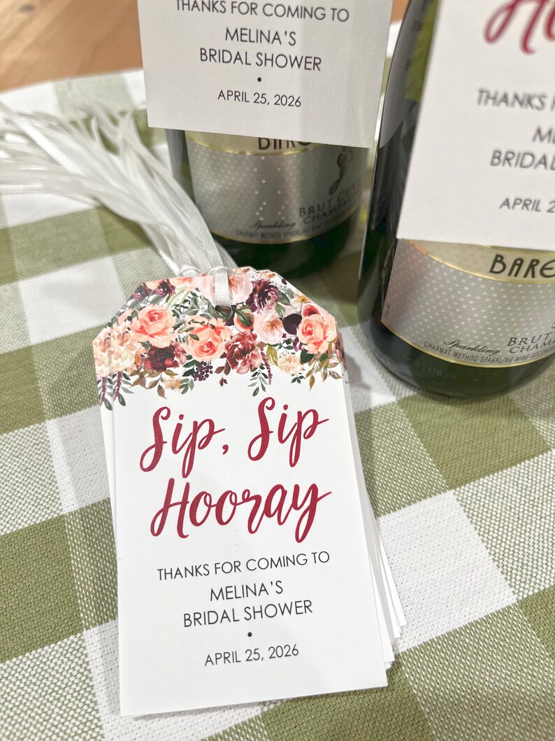 以下が含まれることがあります： 赤い筆記体で「Sip, Sip Hooray」と書かれた白いタグ。タグには「Melina's Bridal Showerへお越しいただきありがとうございます」と「2026年4月25日」と書かれています。背景にはシャンパンボトルがあります。