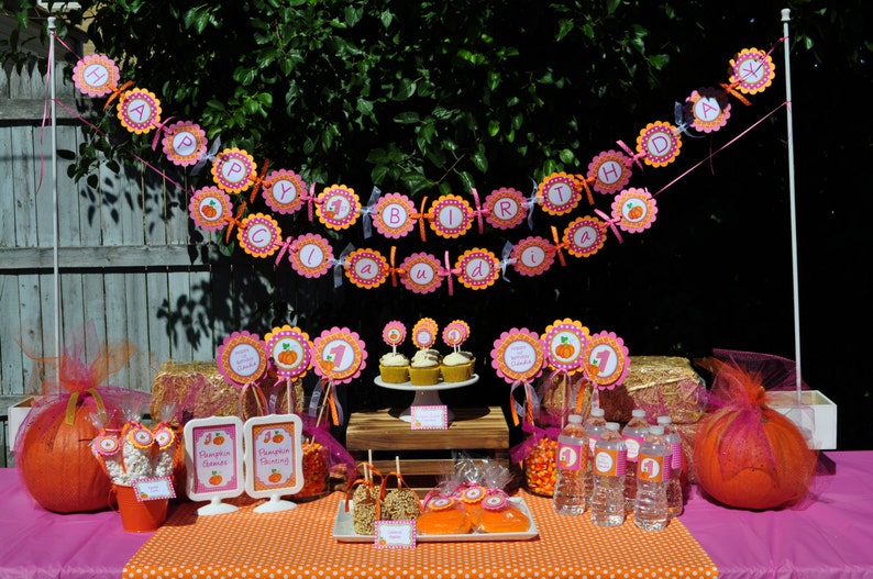May include: A pink and orange pumpkin-themed first birthday party table setting with cupcakes, a banner that says "Happy 1st Birthday", and a variety of decorations.