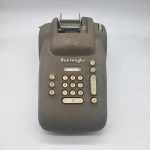 May include: An antique Burroughs adding machine in a dark gray color. The machine has a series of number keys, function keys, and a display window. The word "Burroughs" is printed on the front.