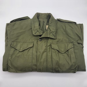 May include: An olive green military-style jacket with a button closure and two front pockets. The jacket has a collared neckline and shoulder epaulets. The fabric appears to be a durable, possibly water-resistant material. The jacket is folded, showing its full shape.