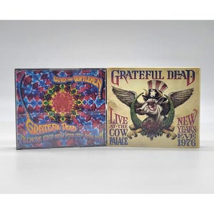 May include: Two Grateful Dead CD cases. One features a colorful, geometric design with the text "Ladies and Gentlemen" and "Fillmore East New York City April 1971." The other has a cow with wings and a top hat, with the text "Live at the Cow Palace New Year's Eve 1976."