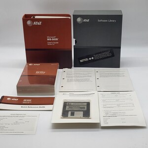 May include: An AT&T Microsoft MS-DOS Version 3.2 software package including a red binder, a grey storage box, a user guide, a quick reference guide, a software registration card, a comment card, and a 5.25-inch floppy disk in a plastic sleeve.