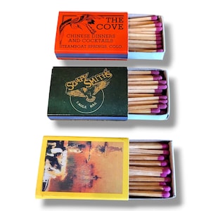 Puede incluir: Tres cajas de cerillas con cajones abiertos, cada una llena de cerillas de madera. La caja superior es naranja con el texto "THE COVE" y "CHINESE DINNERS AND COCKTAILS". La caja del medio es verde con un gráfico de águila y el texto "SOAPY SMITHS". La caja inferior es amarilla.