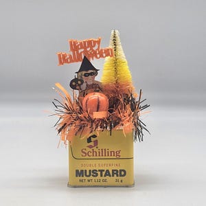 Könnte beinhalten: Eine gelbe Vintage-Senfdose von Schilling, verziert mit Halloween-Dekoration, darunter ein Kinderausschnitt mit Hexenhut, ein orangefarbener Kürbis, ein gelb-oranger Flaschenbürstenbaum sowie orangefarbenes und schwarzes Lametta.