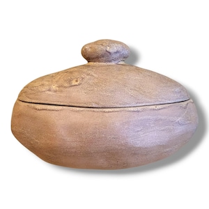 May include: A handcrafted, oval-shaped ceramic container with a lid. The container and lid are a muted, speckled brown color with a textured surface. The lid has a small, rounded knob for easy handling. The container is likely used for storage.