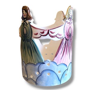 May include: A ceramic candle holder with two figures in long dresses, one green and one pink, holding a banner. The base is decorated with blue cloud-like shapes, and the design includes cut-out holes for light.