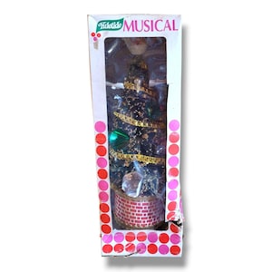 May include: A miniature Christmas tree in a box. The tree is dark green with gold garland and ornaments. The base is a red brick design. The box has the words "Yuletide MUSICAL" printed on it.