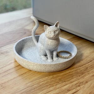 May include: A speckled gray cat-shaped trinket dish with a ring inside. The cat figurine sits in the center of the dish, tail curved upwards. The dish is round and has a speckled pattern.