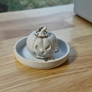May include: A speckled, light gray pumpkin-shaped ring holder with carved facial features, sitting in a matching round dish. Gold rings are placed on the pumpkin and in the dish. The ring holder is on a wooden surface.