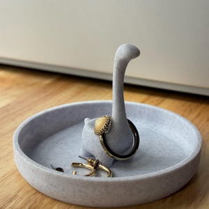 Puede incluir: Un porta anillos de cerámica blanca con forma de dinosaurio de cuello largo. El porta anillos tiene un pequeño plato redondo y un anillo está colocado en el cuello del dinosaurio. También hay dos pequeños pendientes en el plato.